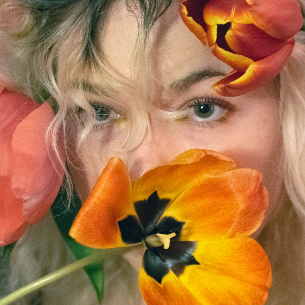 Eyes level with the lens above an open orange tulip that masks nose and mouth, blonde curls and a red bloom behind