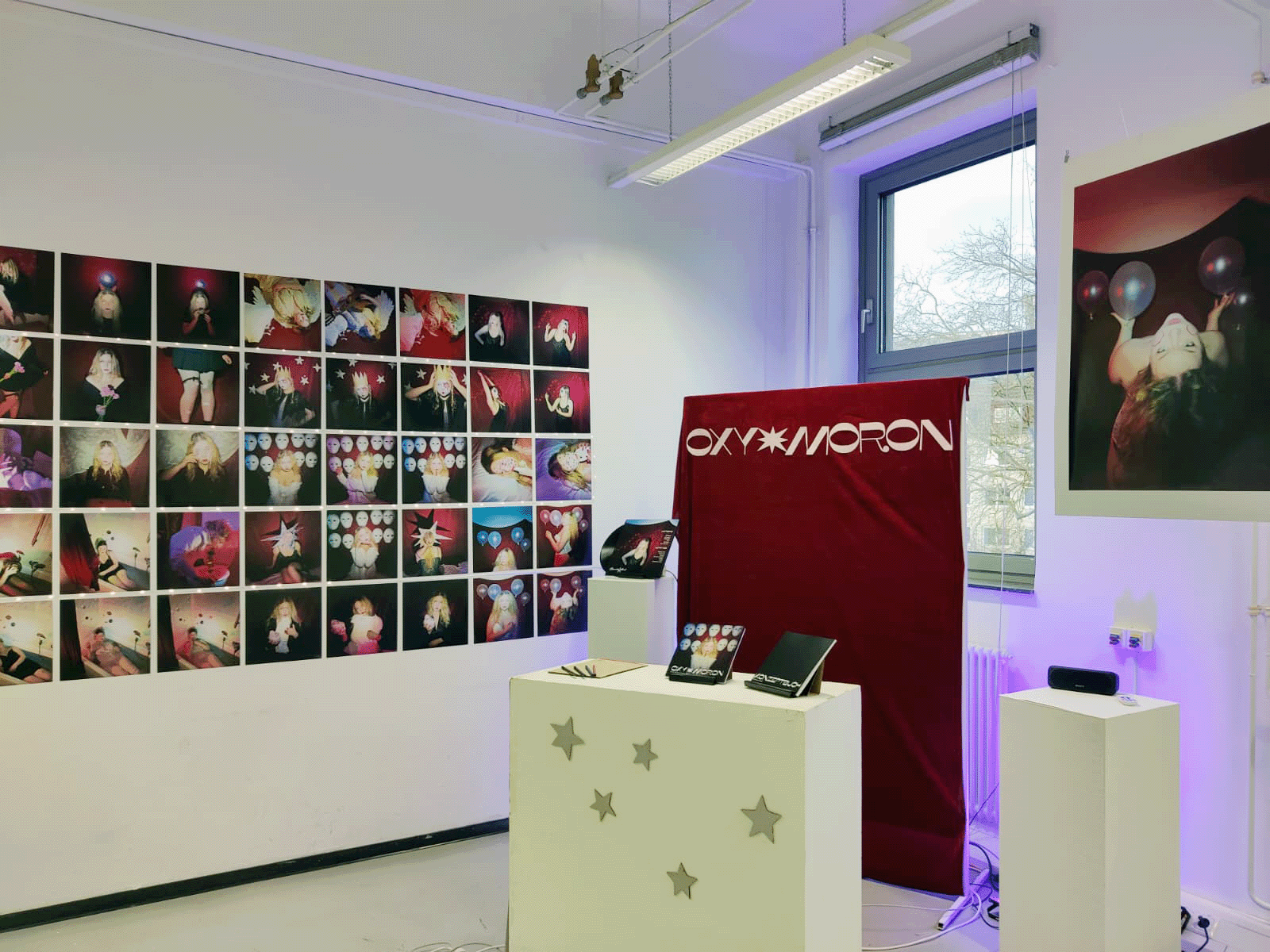 Exhibition view: grid of prints on a white wall, red OXY MORON banner and booklets on white plinths