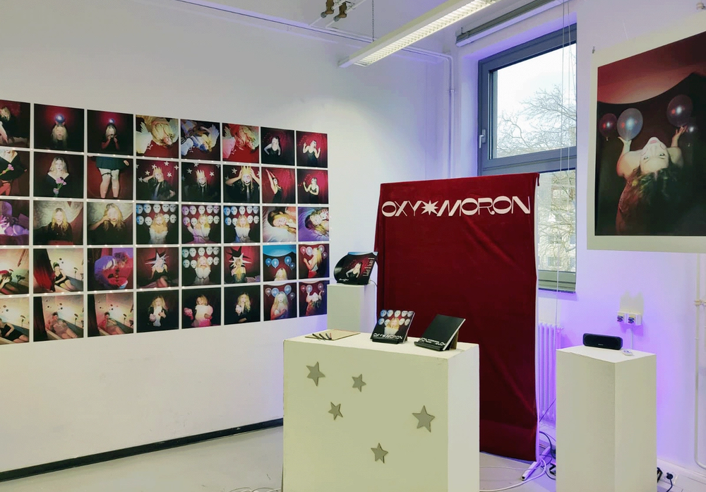 Exhibition view: grid of prints on a white wall, red OXY MORON banner and booklets on white plinths