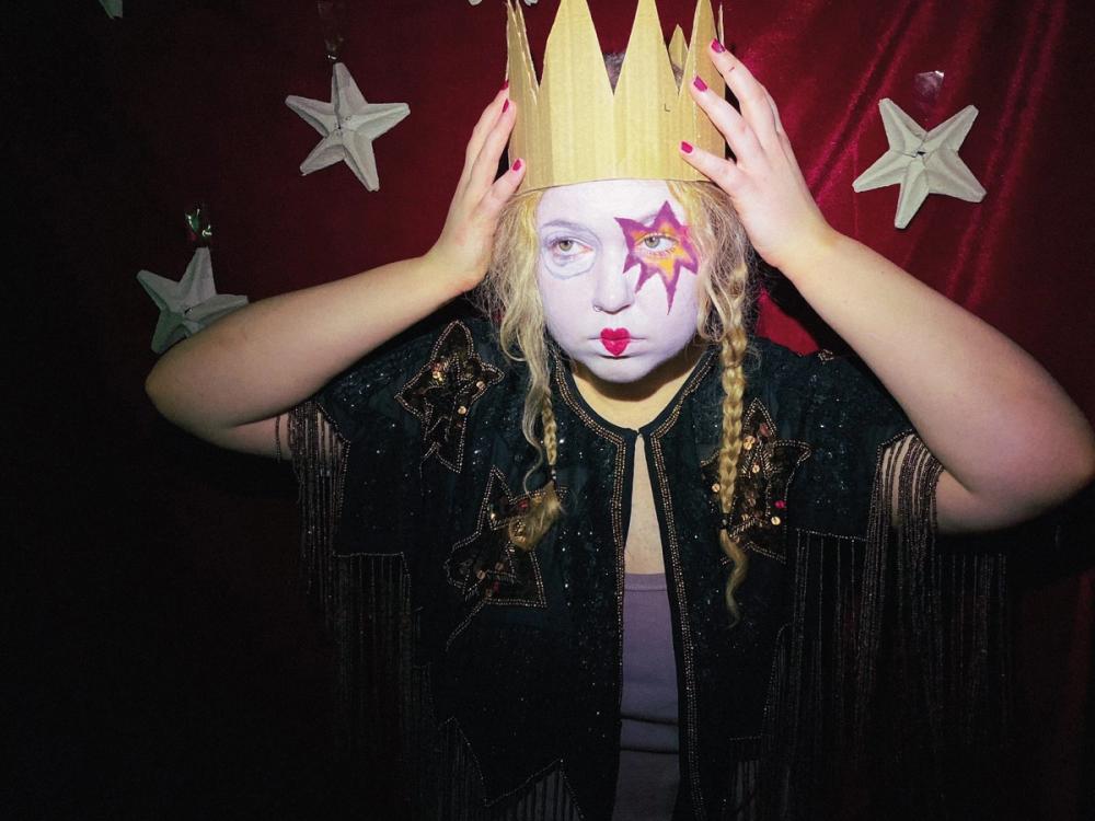 White-painted face with a star around one eye, gold paper crown held up, black fringed sequin jacket
