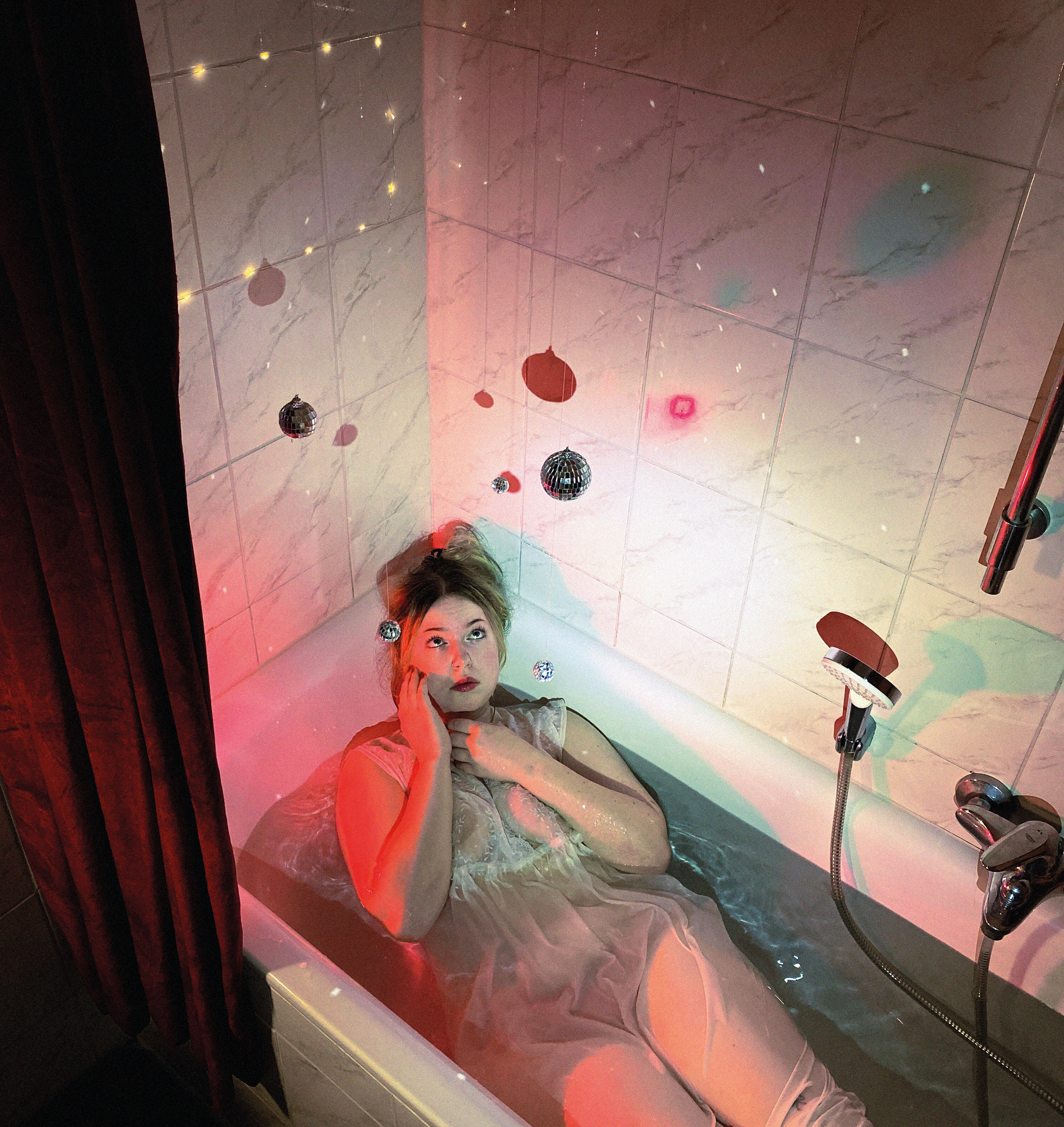 In a bathtub in a sheer dress, disco balls and fairy lights hung across marble tiles under pink light
