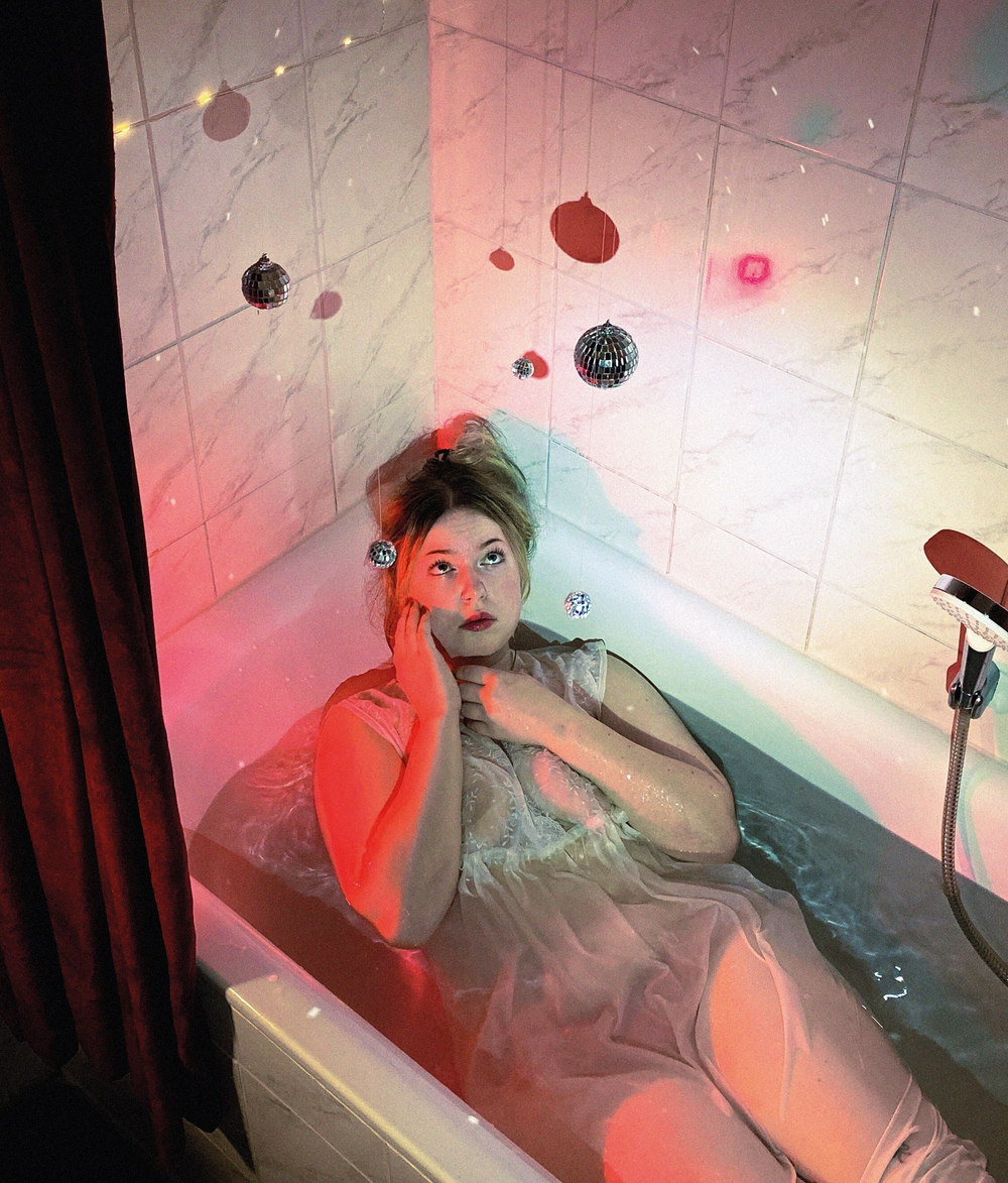 In a bathtub in a sheer dress, disco balls and fairy lights hung across marble tiles under pink light