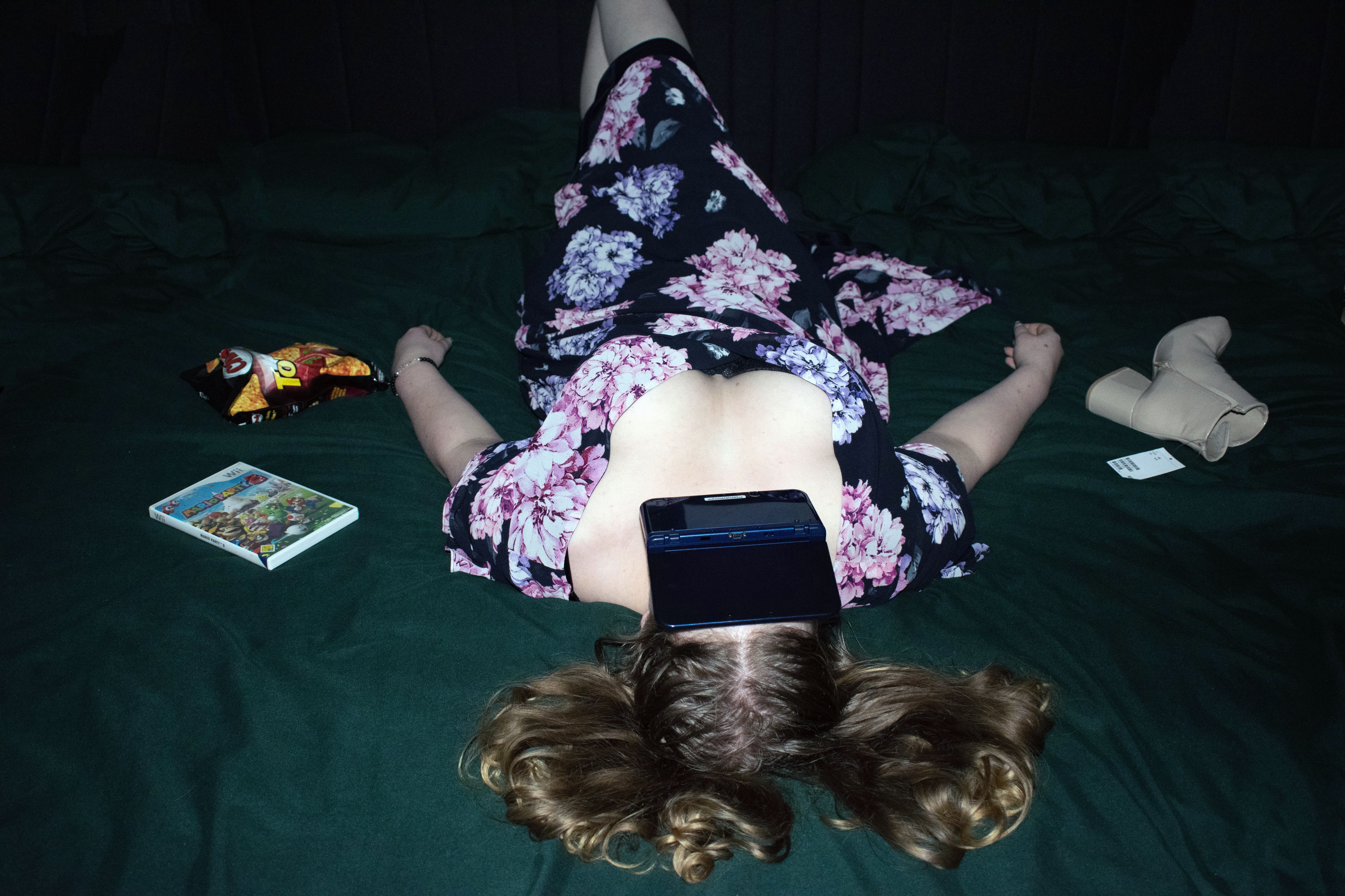 Woman sprawled face-up on a dark green bed, a games console over her face, crisps and boots beside her