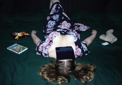 Woman sprawled face-up on a dark green bed, a games console over her face, crisps and boots beside her