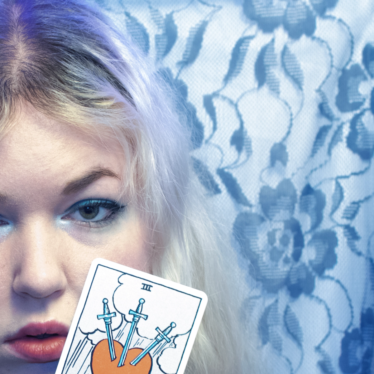 Half a face in cold blue light against floral lace, holding up a tarot card of three swords piercing a heart