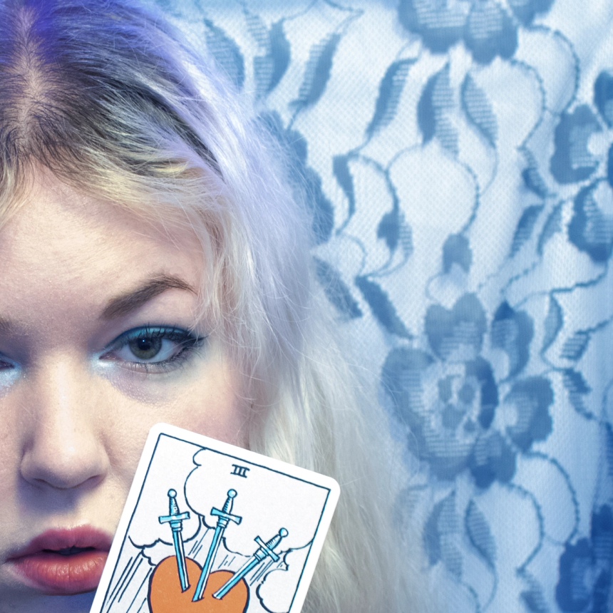 Half a face in cold blue light against floral lace, holding up a tarot card of three swords piercing a heart