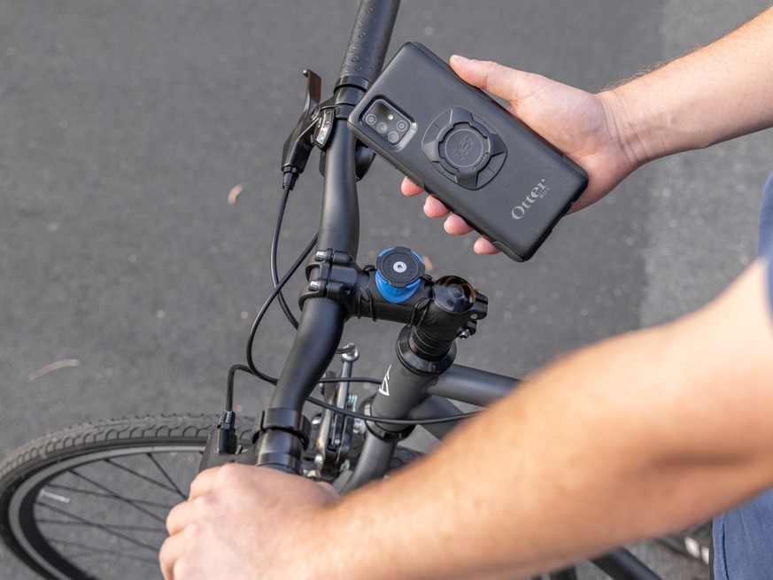 A person holds a smartphone with an OtterBox case over the handlebars of a bicycle, demonstrating a Quad Lock mounting system attached to the bike.