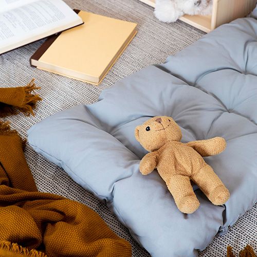 Un osito de peluche marrón claro acostado sobre un cojín de suelo acolchado gris claro. Junto a ellos, hay libros y una manta tejida de color mostaza.