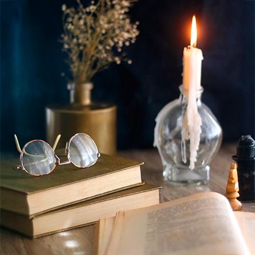 Una mesa con una pila de libros, un par de gafas de montura redonda, un florero con flores secas y una vela encendida goteando cera sobre su base.