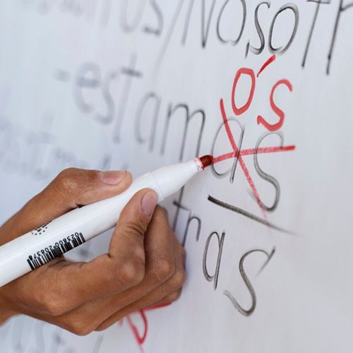 Una mano sosteniendo un marcador rojo sobre una pizarra blanca, tachando la terminación "os" y "as" de la palabra "Nosotros/Nosotras", escribiendo un "ó" con acento.