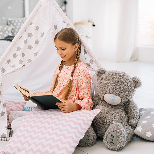 Una niña con trenzas y un vestido estampado de flores, sentada dentro de una pequeña tienda de campaña blanca con estrellas grises. Está leyendo un libro, y un gran oso de peluche gris está sentado a su lado.