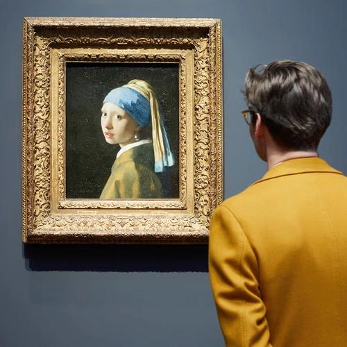 Un hombre con una chaqueta amarilla y gafas mirando el cuadro "La joven de la perla" de Vermeer en un museo.