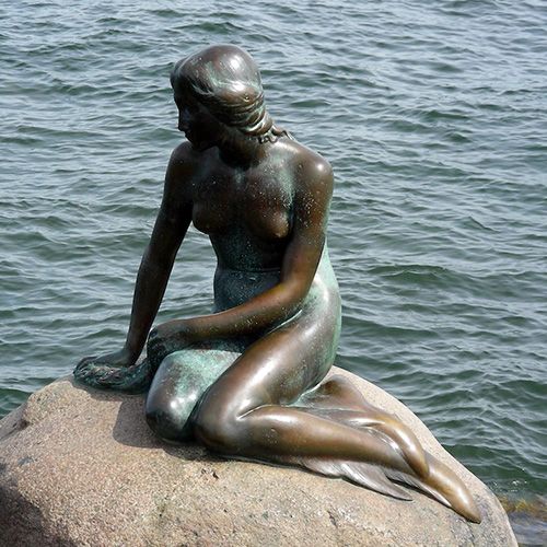 Una estatua de bronce de la Sirenita sentada sobre una roca, con el mar azul de fondo.