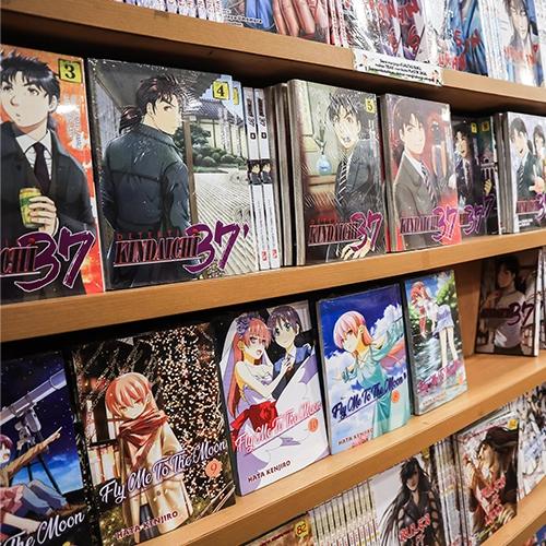 Un estante de tienda lleno de volúmenes de manga, incluyendo "Detective Kindaichi 37" y "Fly Me To The Moon".