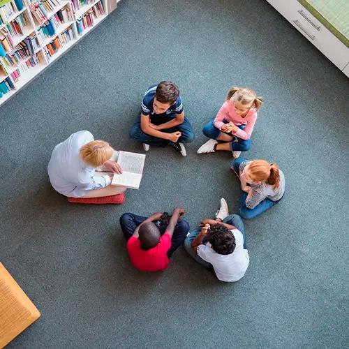 Una vista aérea de cinco niños y un adulto sentados en círculo en el suelo de una biblioteca, con el adulto leyendo un libro.