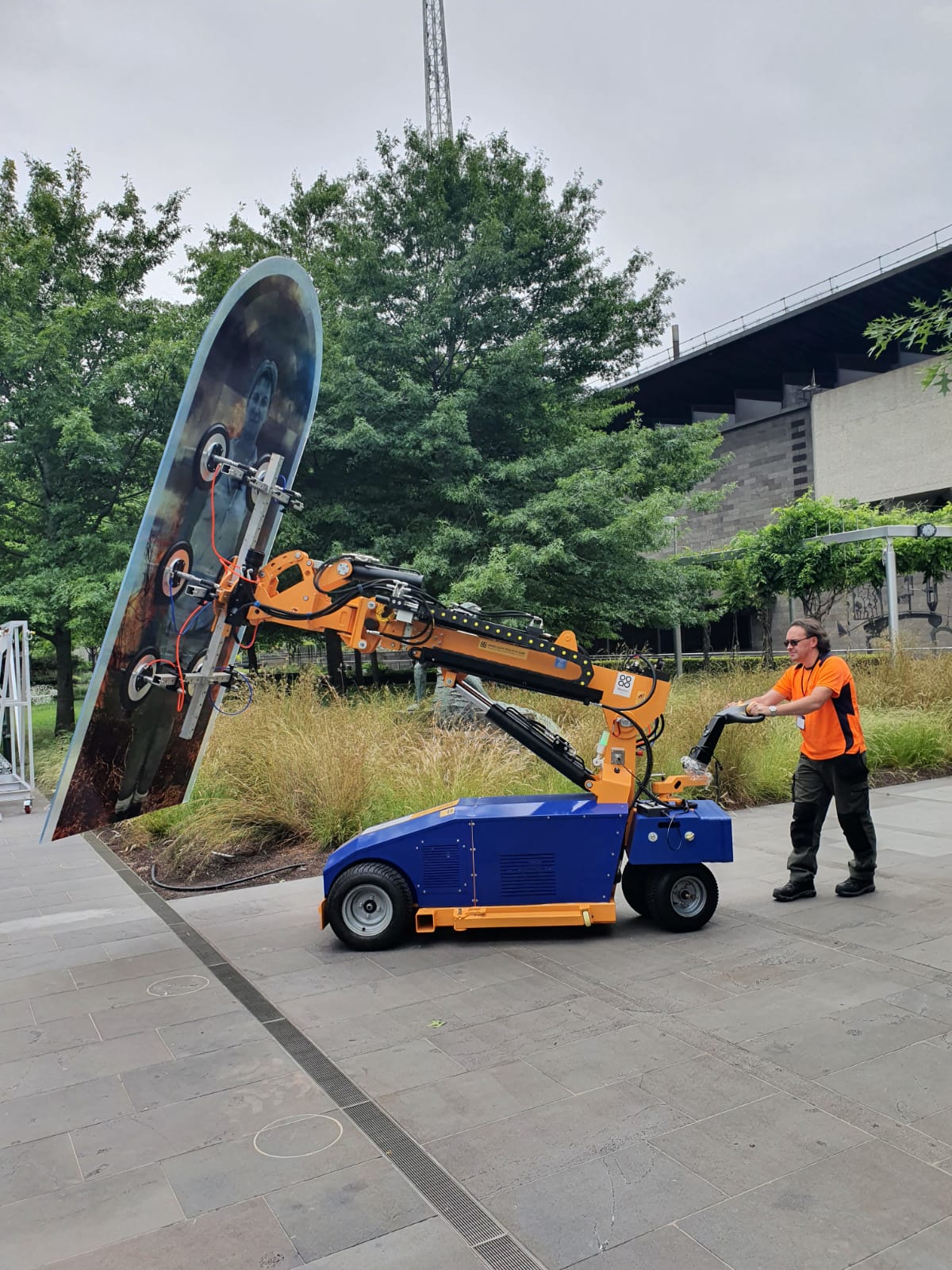 Quattrolifts powered glass lifter holding a tall printed glass art panel outside a gallery building