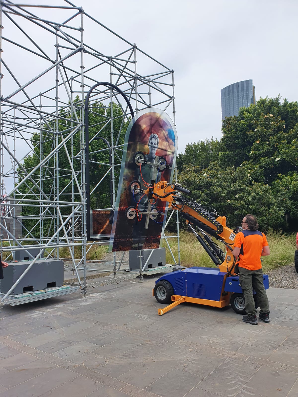 Quattrolifts crawler vacuum lifter holding a tall printed glass panel into a museum exhibit frame outdoors