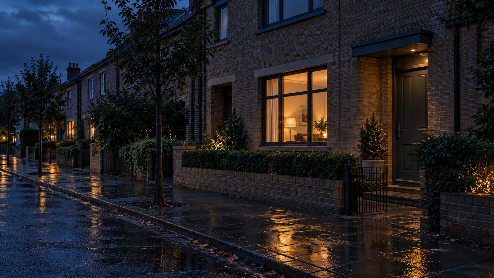 A warmly lit window in a brick townhouse overlooks wet pavement at blue hour.