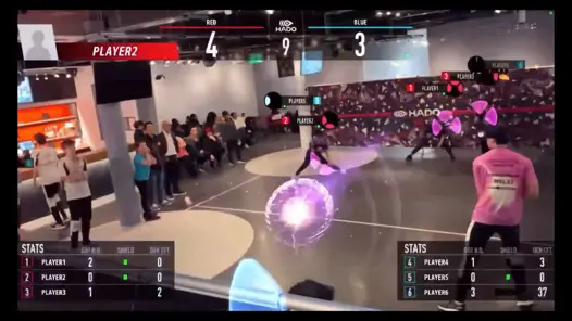A HADO match with players wearing headsets, launching virtual energy balls while defending with shields. The score is Red: 4, Blue: 3, with stats displayed and spectators watching.