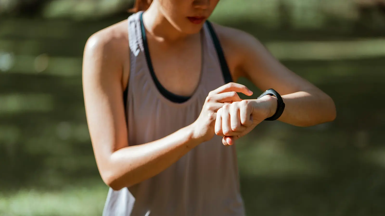 A woman in athletic wear checking her smartwatch while standing outdoors, surrounded by greenery and soft, natural light.