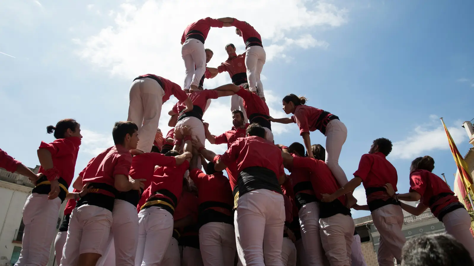 A human tower (castell) formed by people in red shirts and white pants, balancing on each other’s shoulders under a sunny sky.