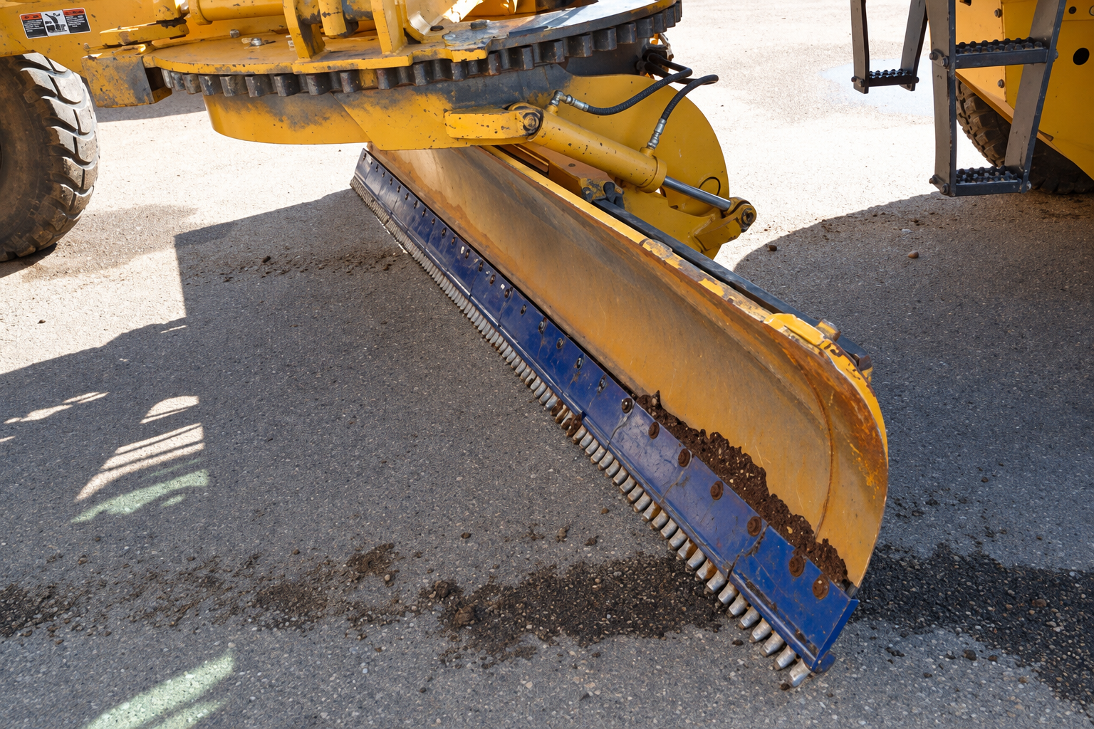 Close-up of a yellow motor grader blade fitted with blue YESS cutting-edge boards and carbide teeth on an asphalt surface.