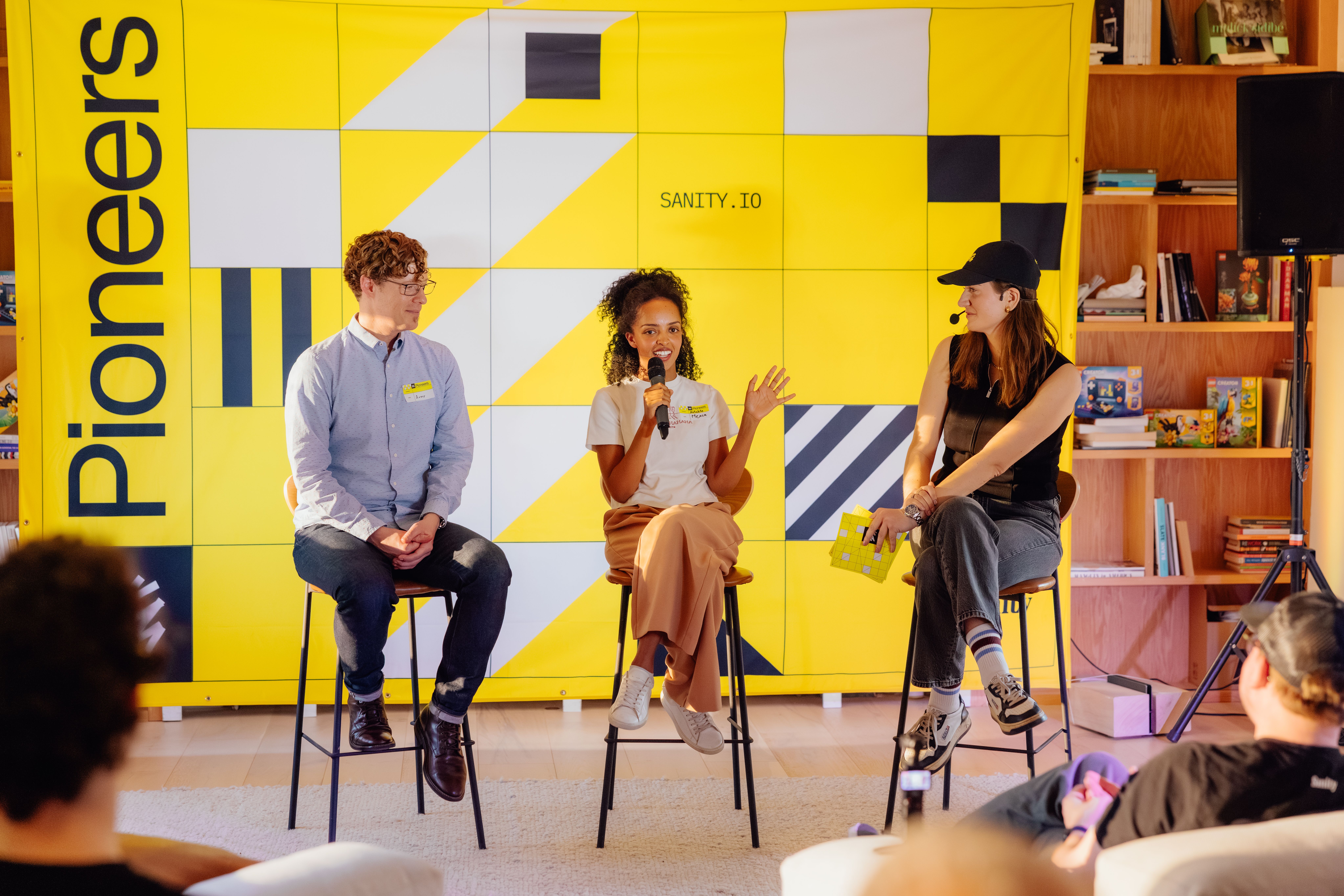 A panel discussion with three people, a man and two women, seated on stools in front of a yellow and black "Pioneers" and "SANITY.IO" backdrop. The woman in the middle is speaking into a microphone.