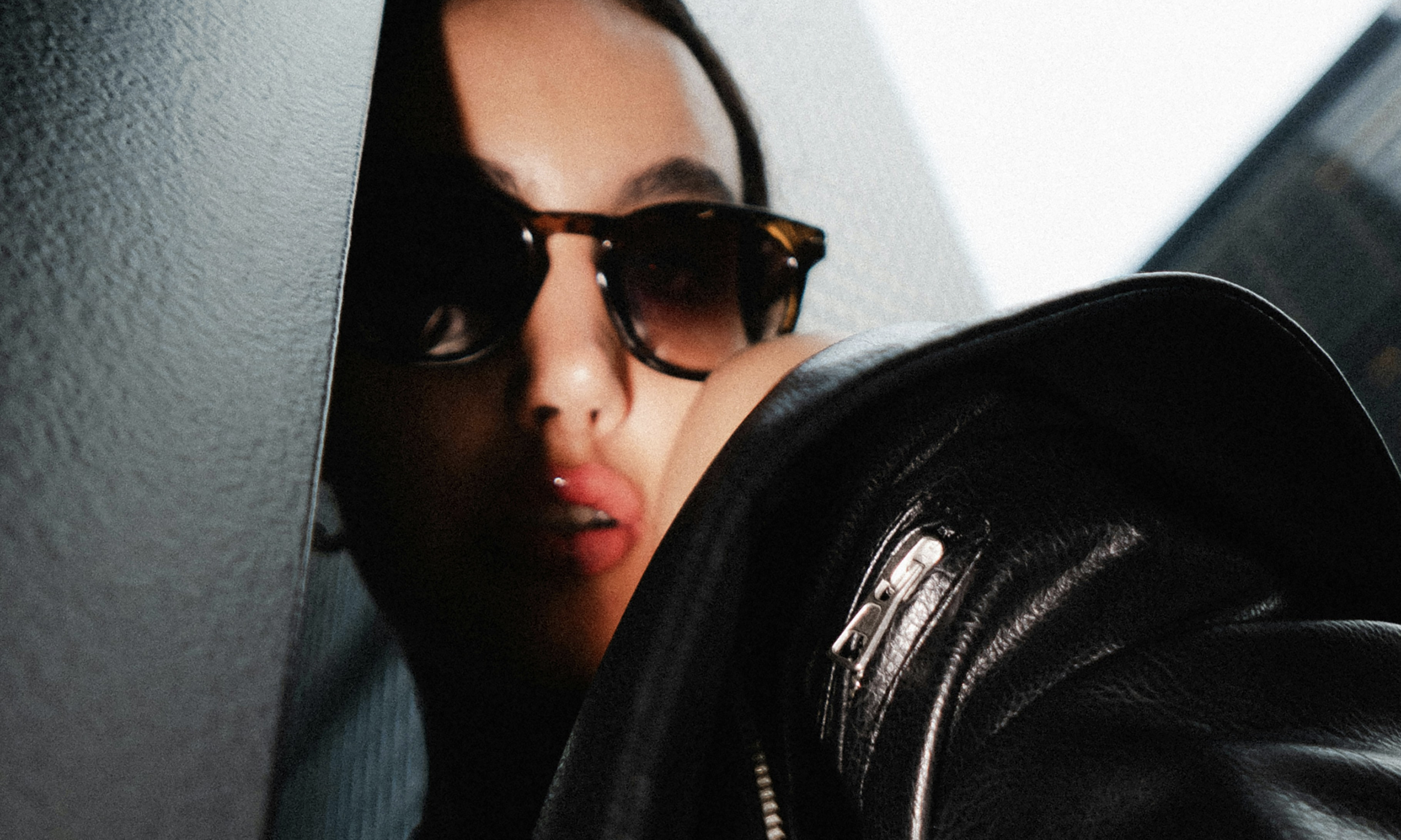 A woman in sunglasses and a black leather jacket with glossy lips, partially obscured by a dark wall.