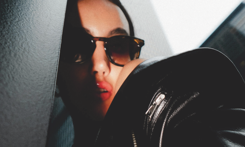 A woman in sunglasses and a black leather jacket with glossy lips, partially obscured by a dark wall.