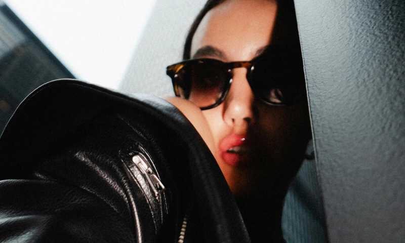 A woman in sunglasses and a black leather jacket with glossy lips, partially obscured by a dark wall.