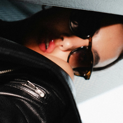 A woman in sunglasses and a black leather jacket with glossy lips, partially obscured by a dark wall.