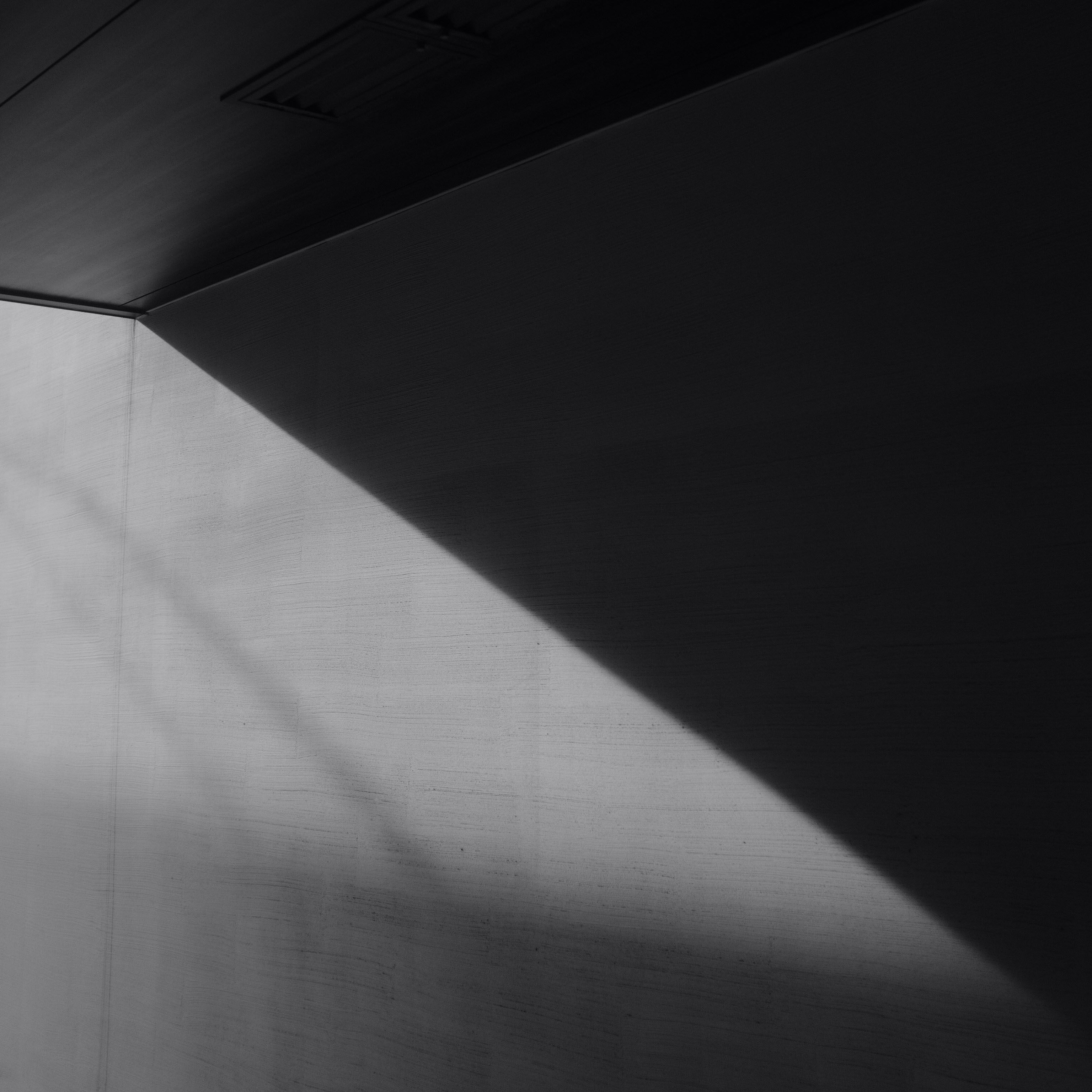 Monochrome image of a room corner, diagonally split by light and shadow, showing wall texture and a ceiling vent.