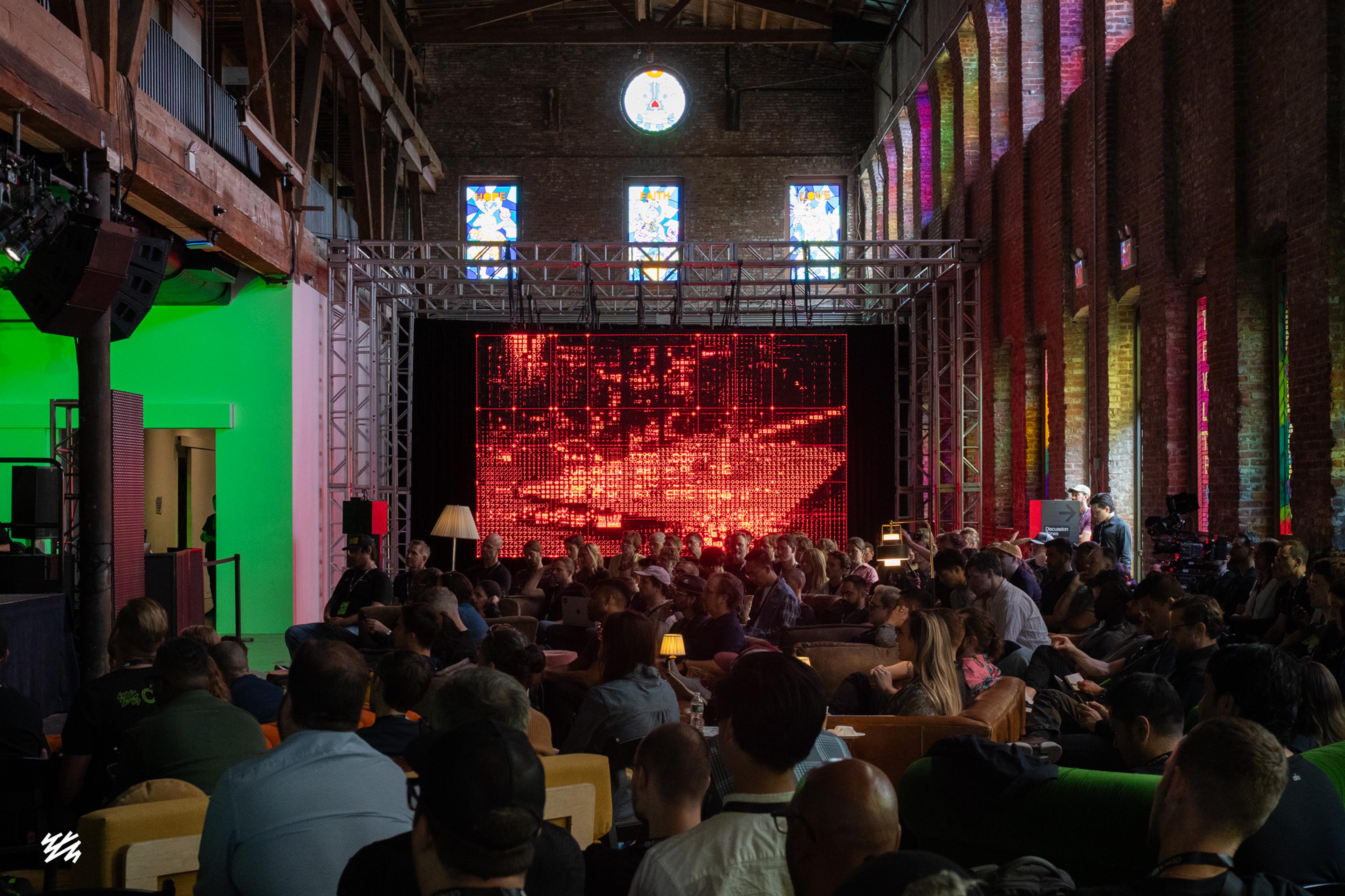 A packed audience at Pioneer Works watches four massive LED screens displaying red text and patterns. The industrial space features exposed brick walls, stained glass windows, and a mix of sofas and chairs. A green-lighted wall is visible on the left side.