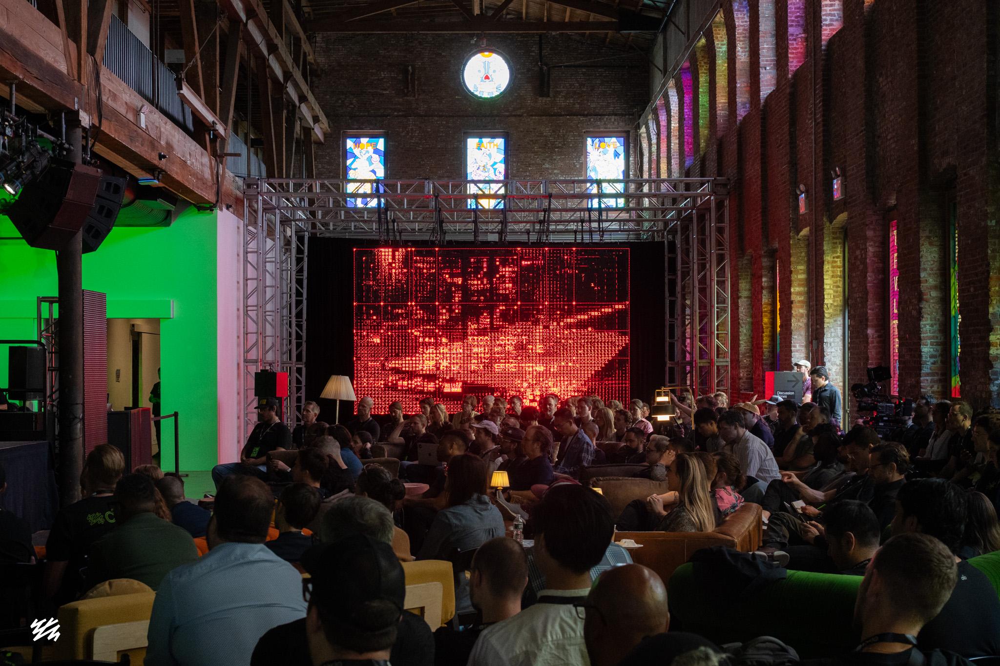 A packed audience at Pioneer Works watches four massive LED screens displaying red text and patterns. The industrial space features exposed brick walls, stained glass windows, and a mix of sofas and chairs. A green-lighted wall is visible on the left side.