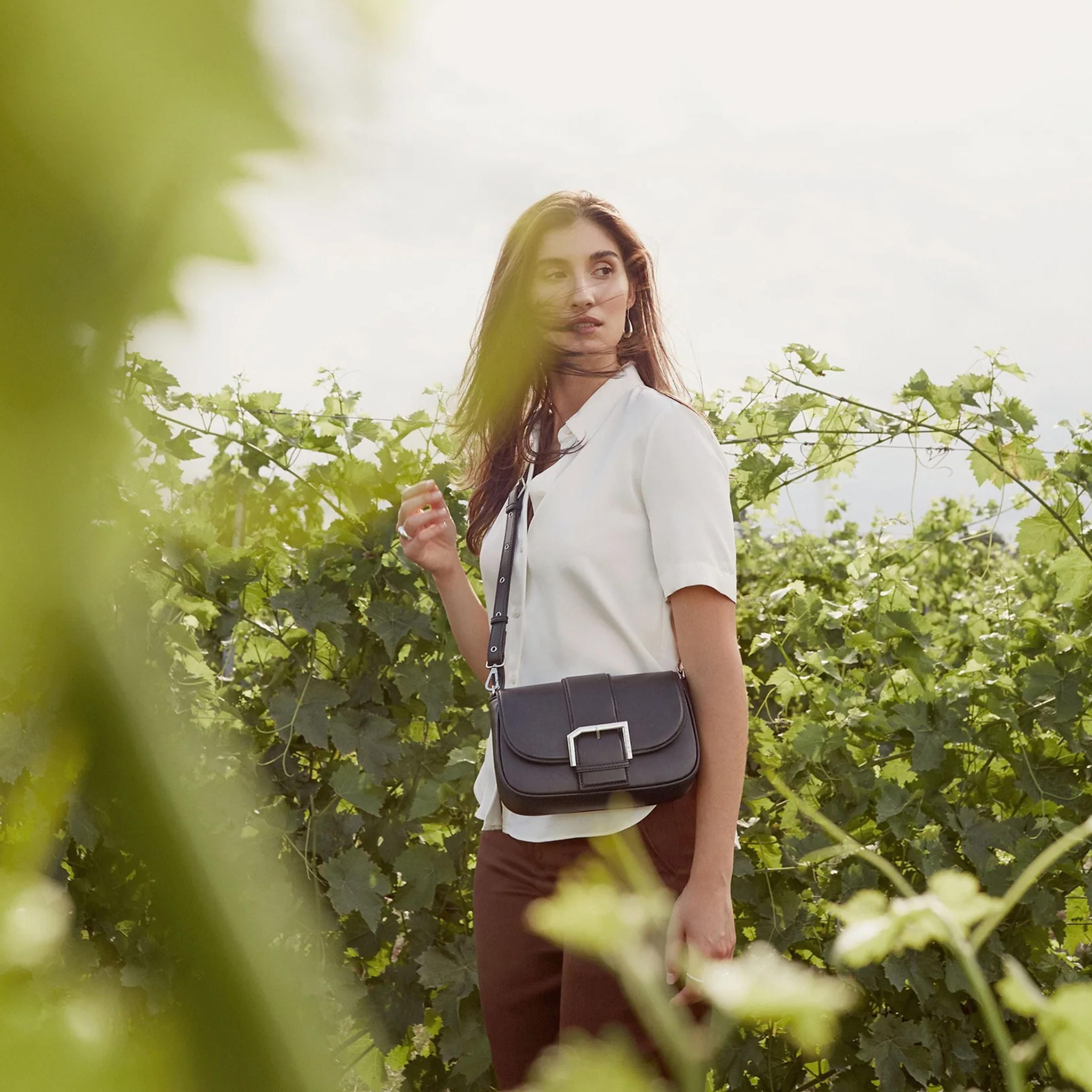 a woman is standing in a vineyard holding a black purse .