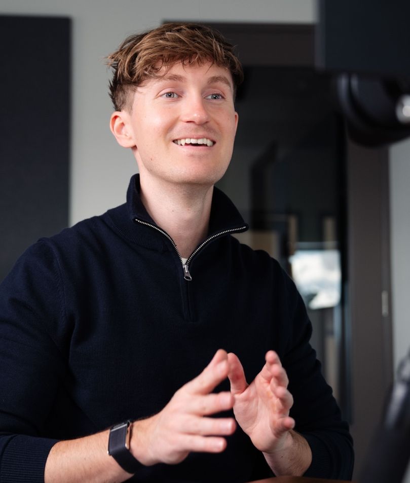 A smiling man in a navy sweater gestures while speaking to a camera.