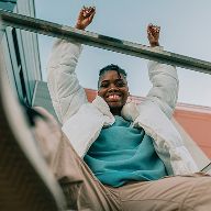 A smiling Black woman in a white puffer jacket holds an overhead bar with raised arms, photographed from a low angle.