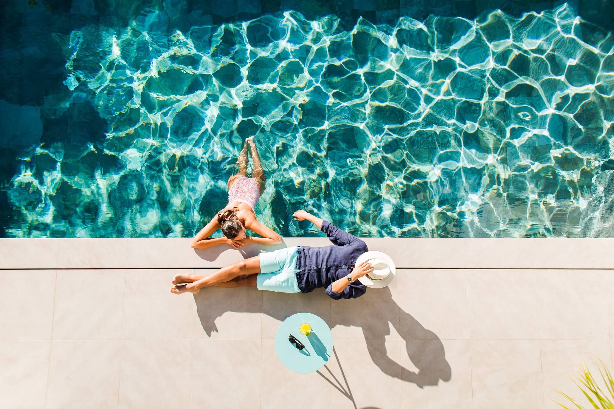 Overhead view of a woman in a blue swimming pool and a man on the sunny deck beside her.