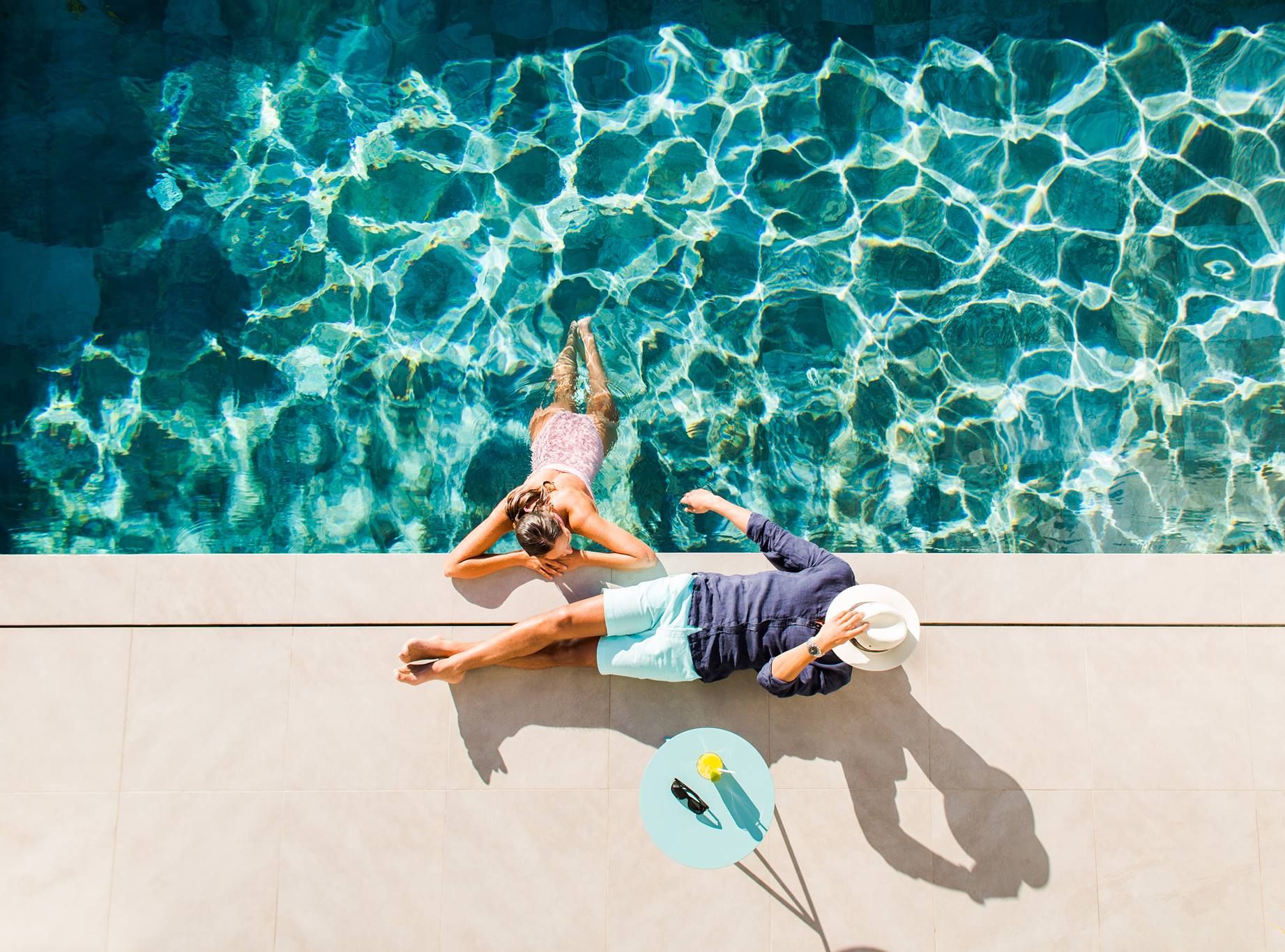 Overhead view of a woman in a blue swimming pool and a man on the sunny deck beside her.