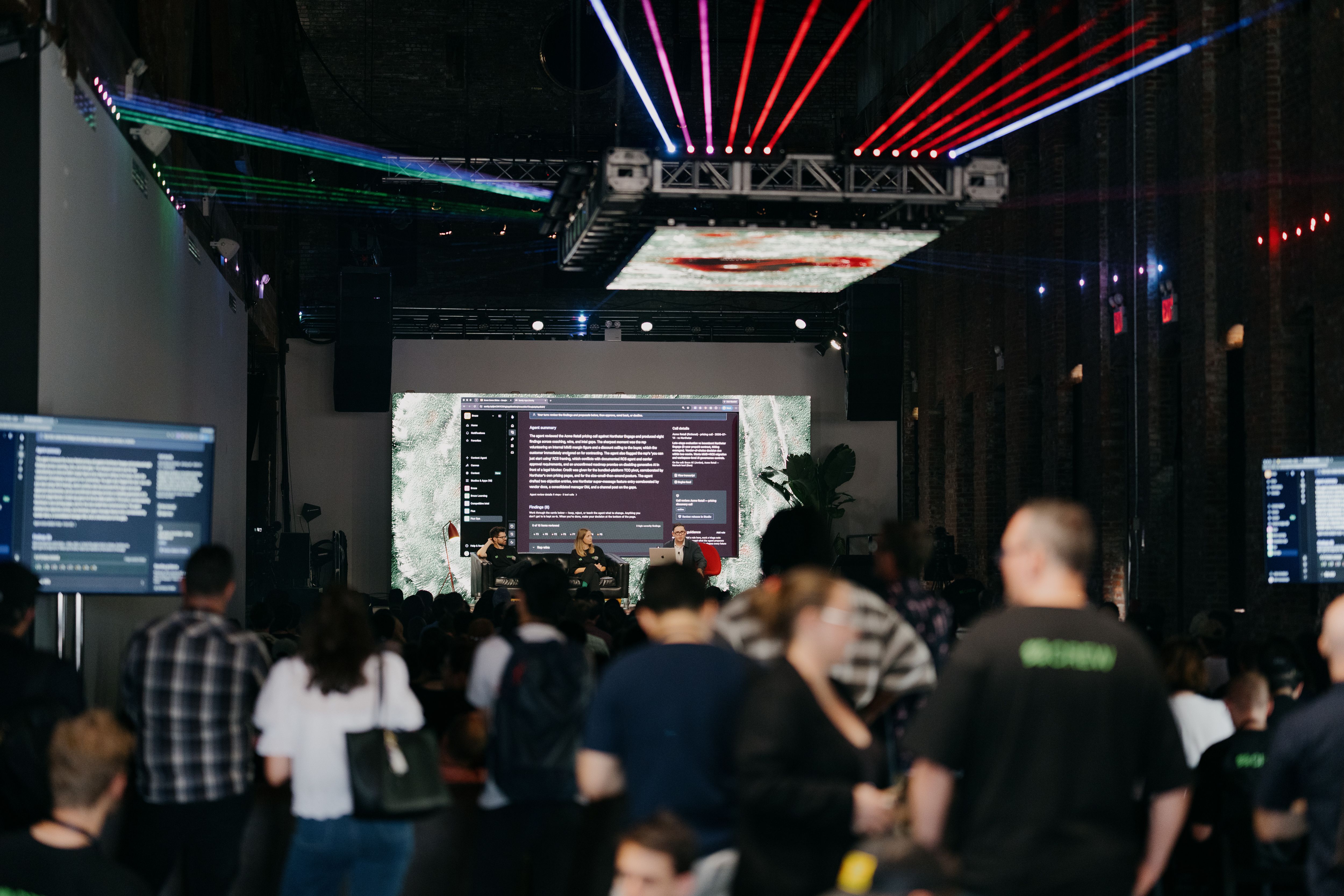 A large audience gathers at an indoor event watching a panel discussion on a stage with multiple screens and colorful lighting.