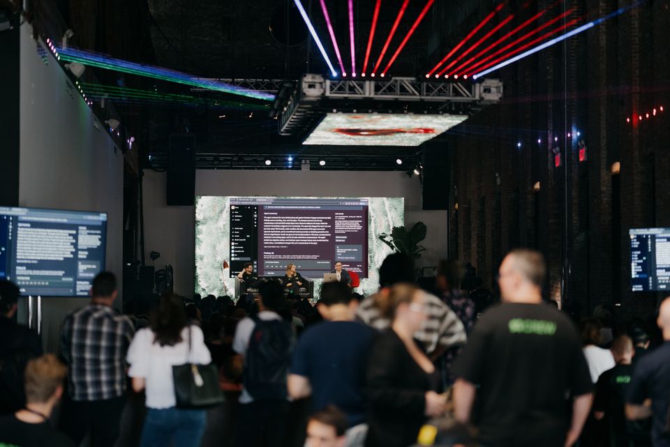 A large audience gathers at an indoor event watching a panel discussion on a stage with multiple screens and colorful lighting. A large audience gathers at an indoor event watching a panel discussion on a stage with multiple screens and colorful lighting.