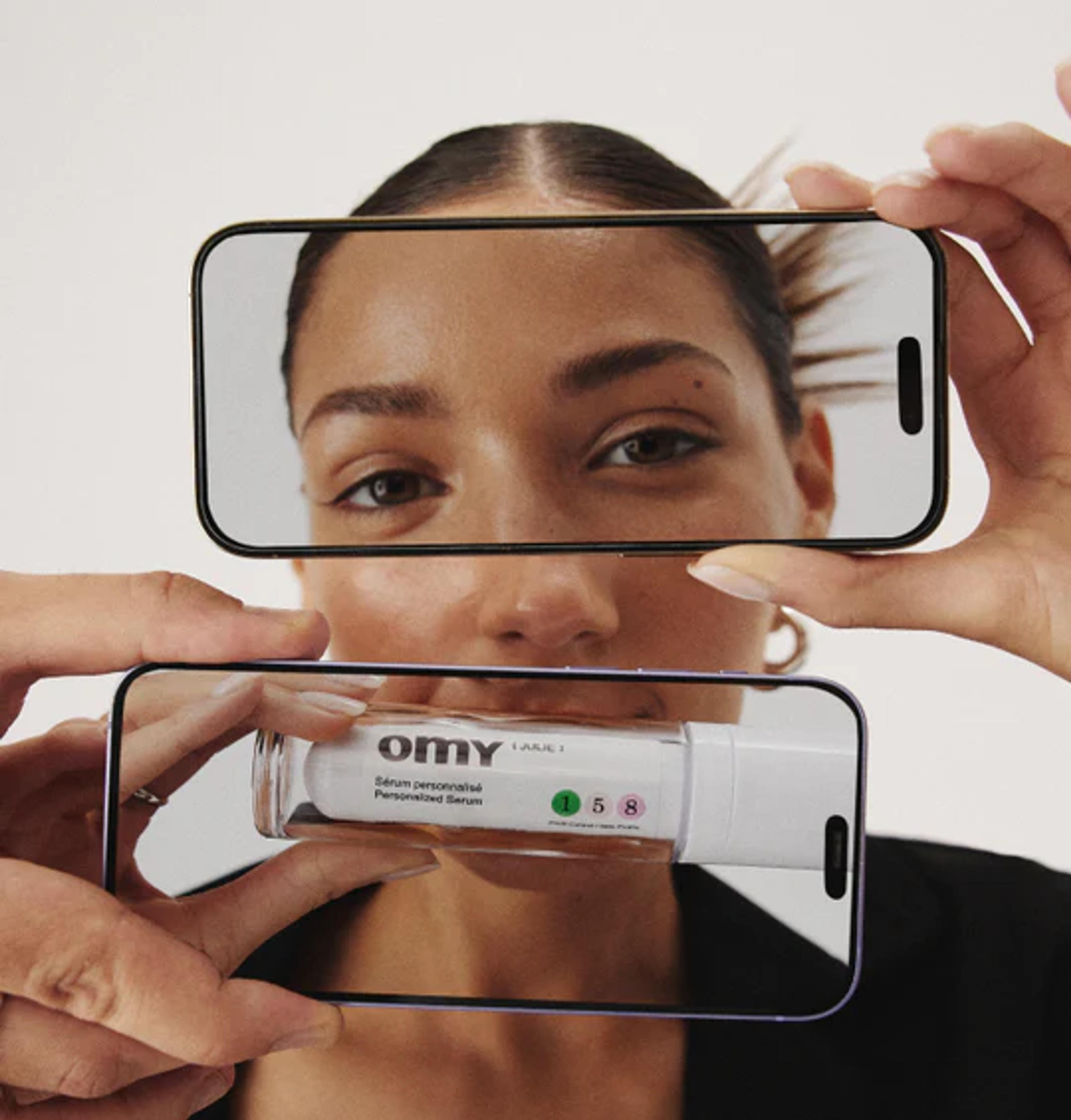 a woman is holding a cell phone in front of her face and a bottle of omy serum
