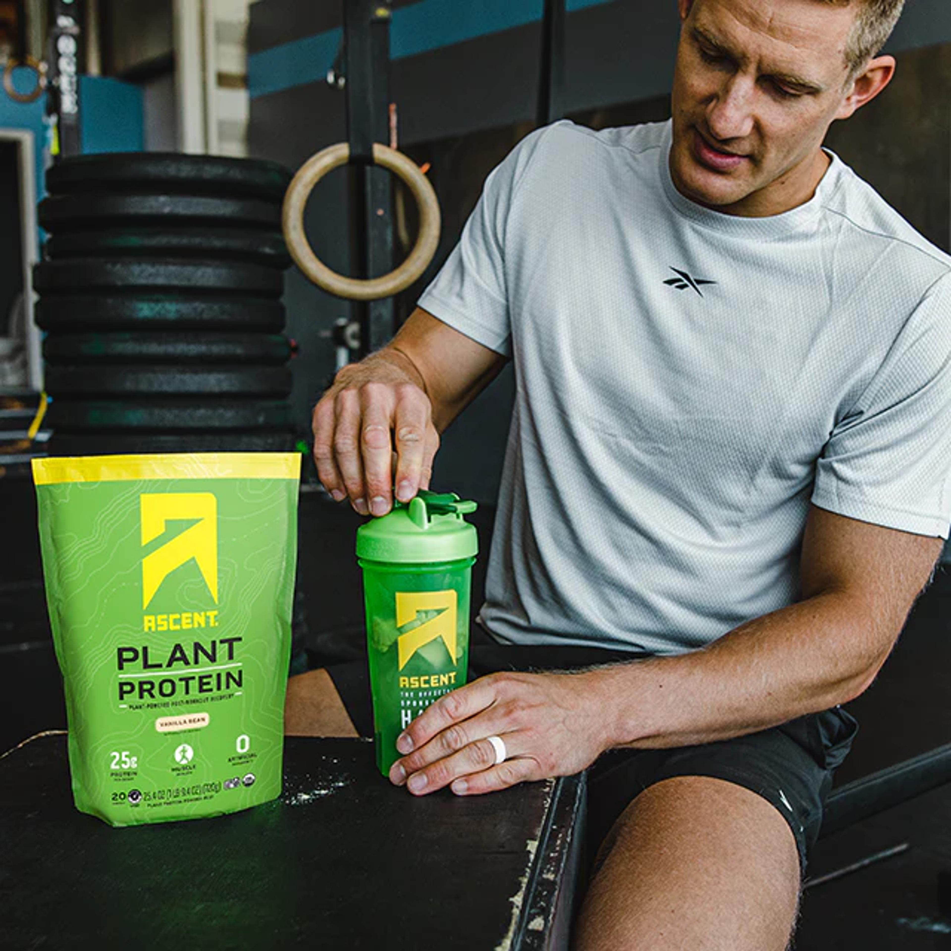 a man is sitting on a bench next to a bag of plant protein and a shaker .
