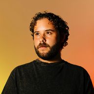 A man with dark curly hair and a beard looks up and to the left against a warm orange-yellow gradient background.