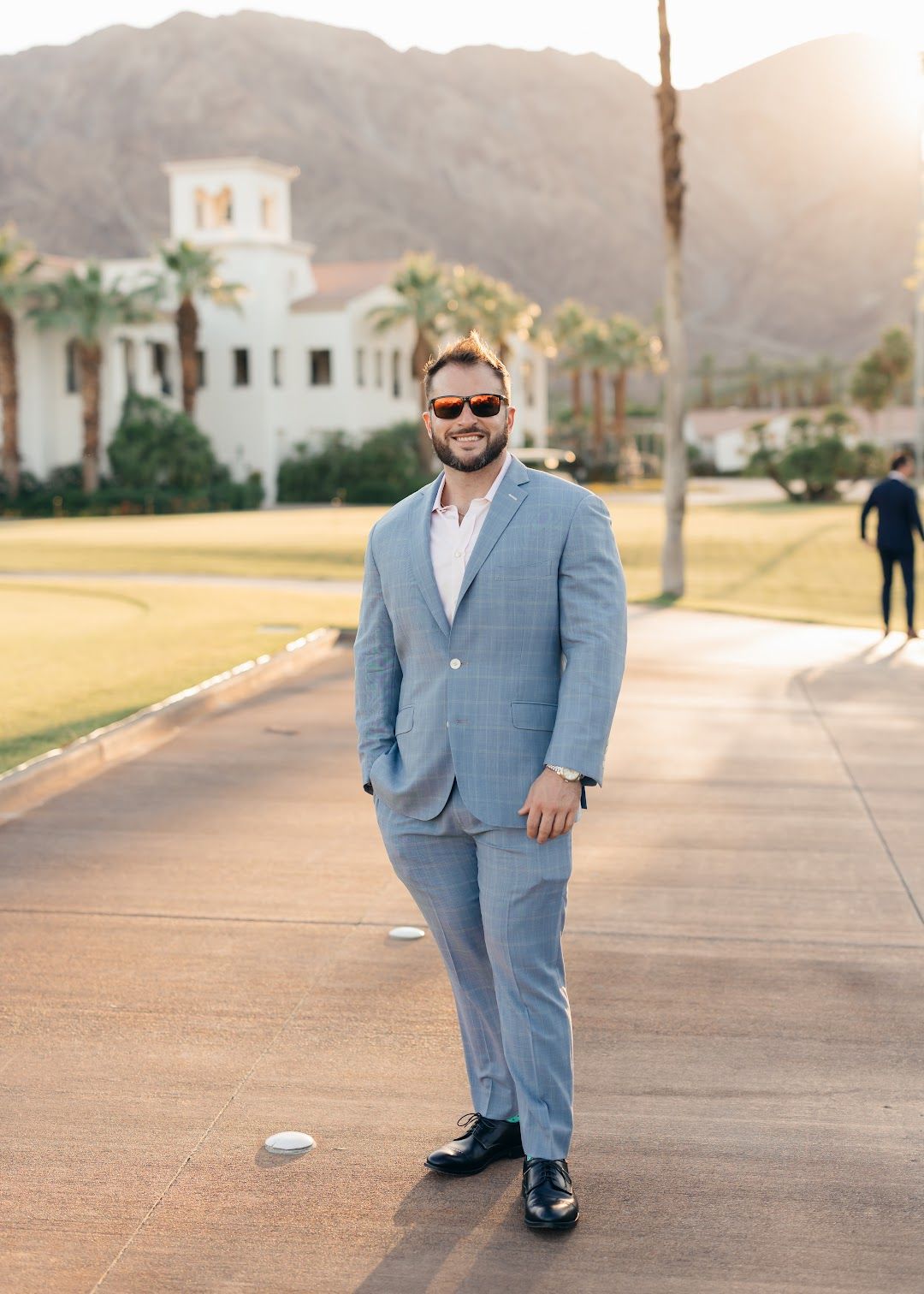 A smiling man in a light blue plaid suit and sunglasses stands on a paved path with a white building and mountains in the background.