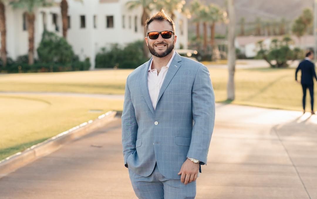 A smiling man in a light blue plaid suit and sunglasses stands on a paved path with a white building and mountains in the background.