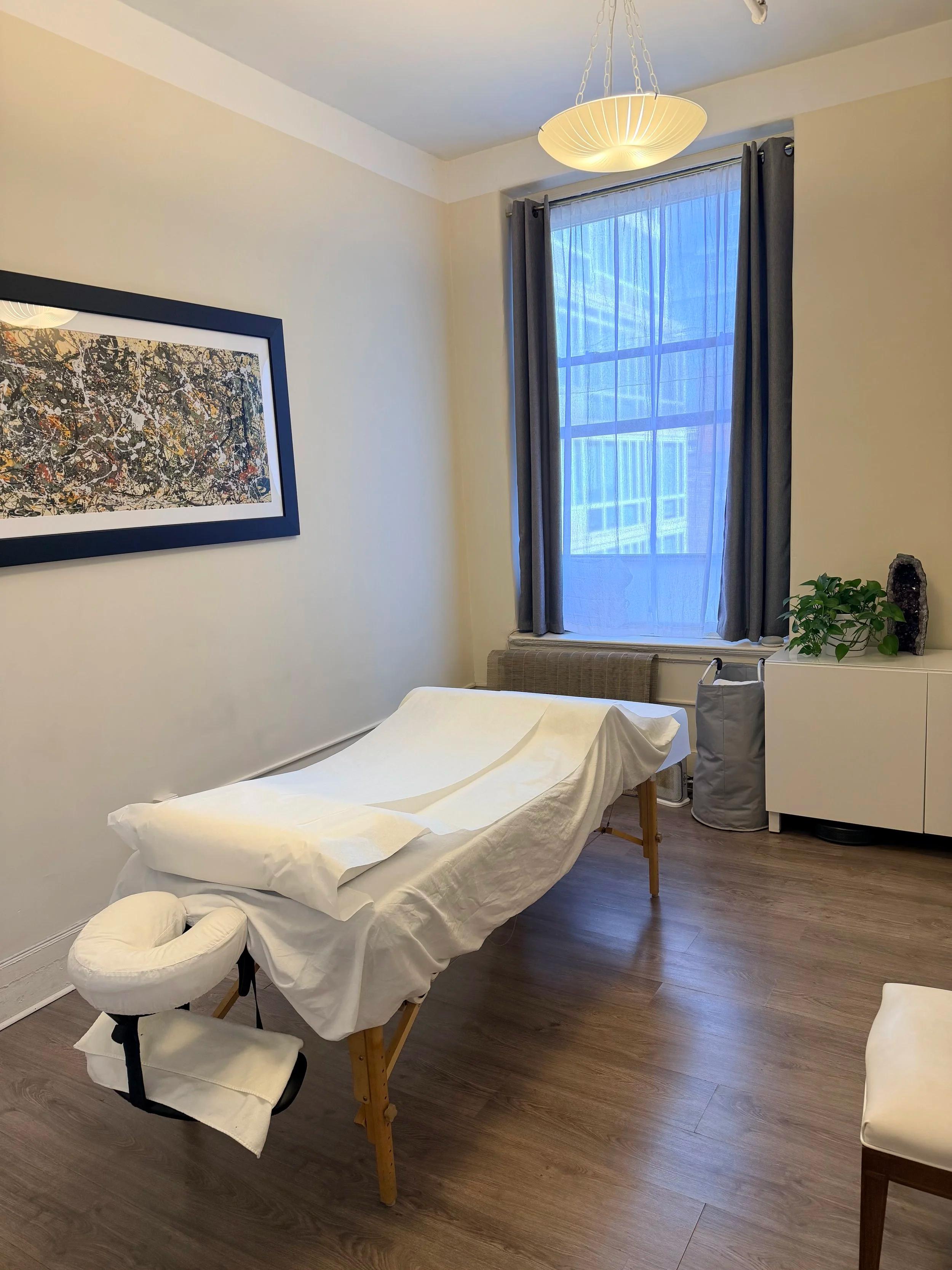 Acupuncture treatment room with a treatment table, window, artwork, and plants.