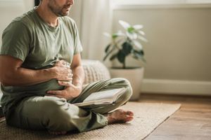 Un uomo seduto a gambe incrociate accanto alla finestra, con le mani sulla pancia.