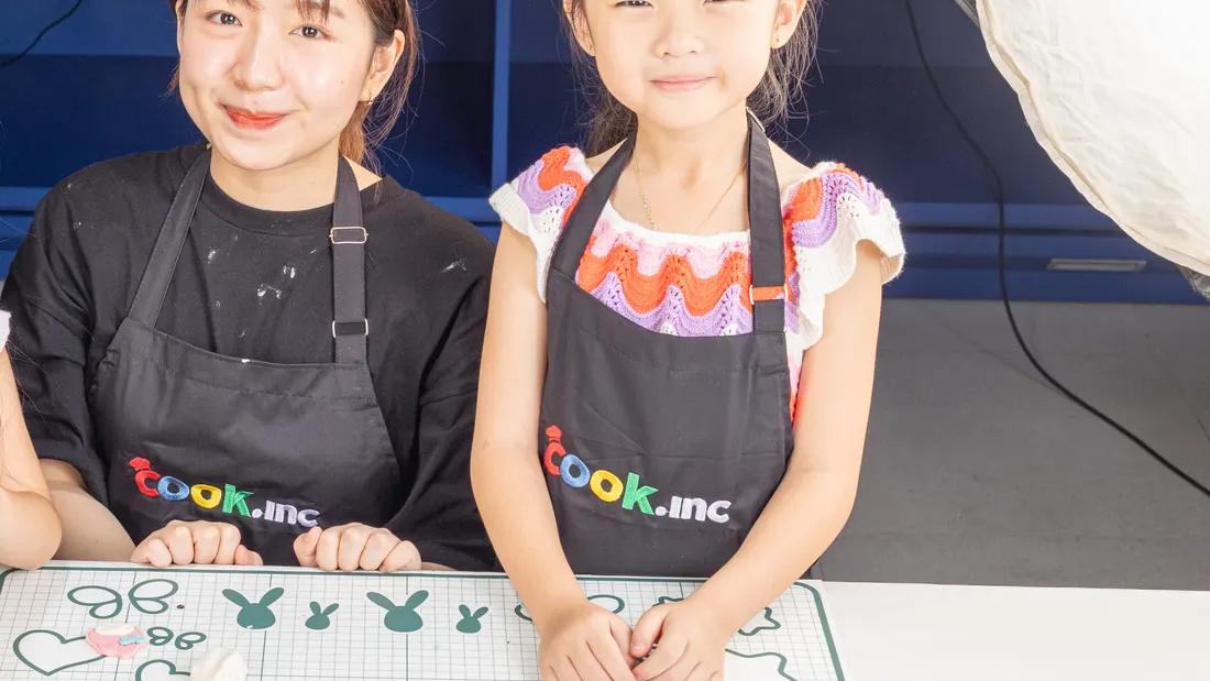 Seorang koki cilik bersama pendamping mengenakan celemek Cook.inc di meja kerja anak