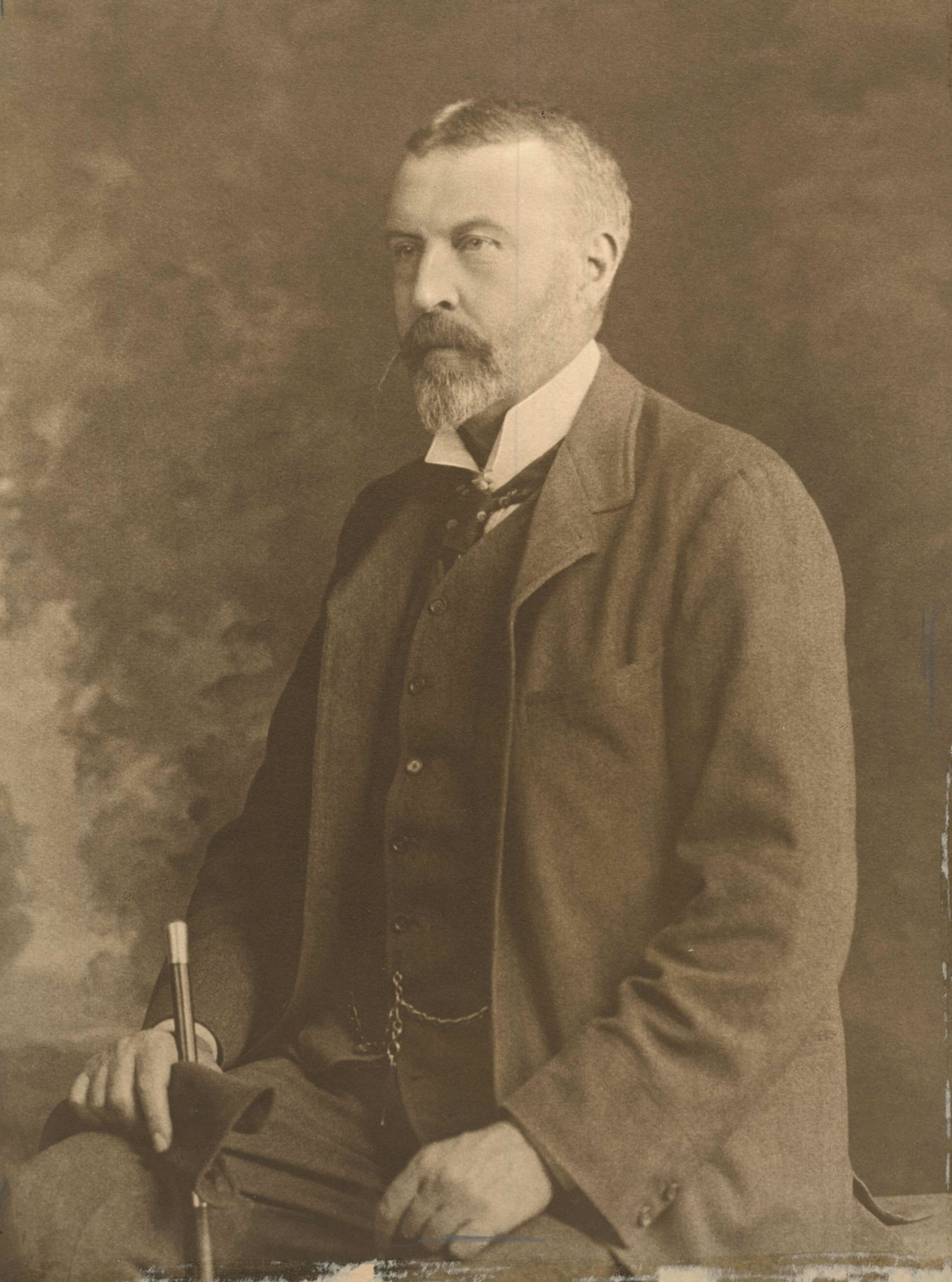 Sepia portrait of John Cathcart Wason, a bearded man in a dark suit, seated and holding a walking cane.